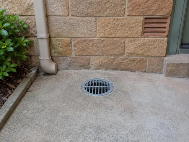Outdoor grated drain set in dry concrete paving beside a brick house wall in Lindfield