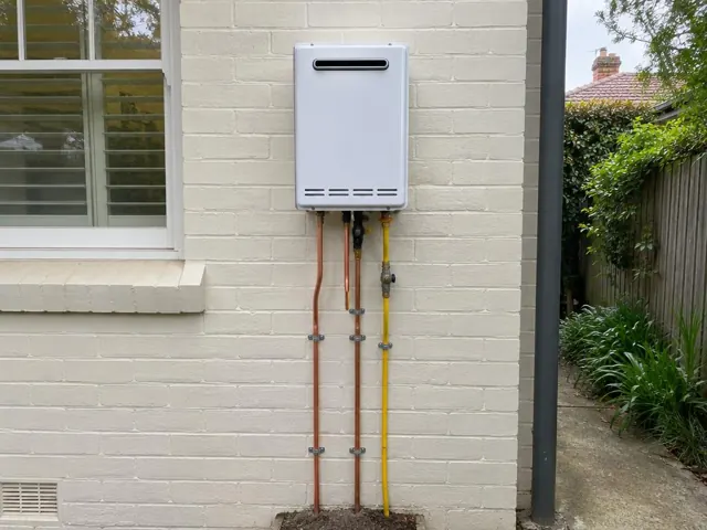 Wall-mounted hot water unit with exposed copper and yellow pipes on a brick house exterior in Pymble